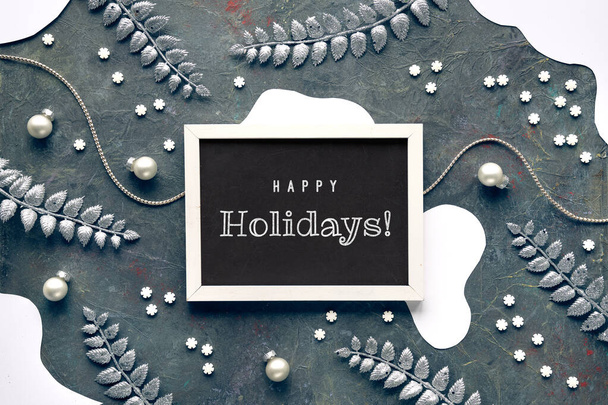 Winter background, text Happy Holidays. Xmas border with fir twigs, Christmas decoration baubles and exotic fern leaves and light garland. Acrylic background in dark grey. Flat lay, top view. - Foto, Imagen