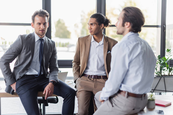 Multikulturelle Geschäftsleute in formeller Kleidung reden, während sie auf Schreibtischen im Büro sitzen - Foto, Bild