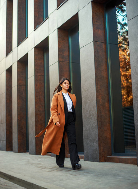 Asian business woman in a long beige coat and hat walks down a modern street on the background of a building in urban style - Foto, imagen