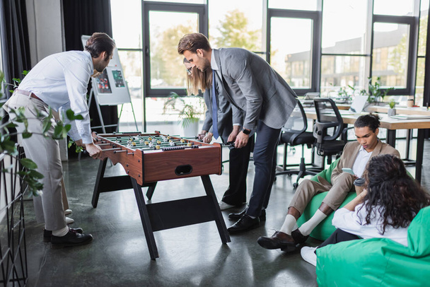 young business people playing table football near multiethnic colleagues sitting in bag chairs - Foto, Bild