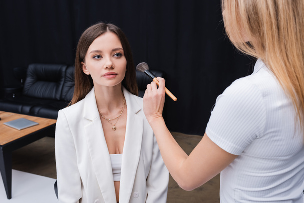 blonde makeup artist holding cosmetic brush near young professional model - Fotoğraf, Görsel