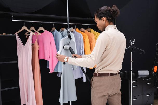Vestido de planchar afroamericano con vaporizador cerca de rack con ropa en vestidor - Foto, imagen