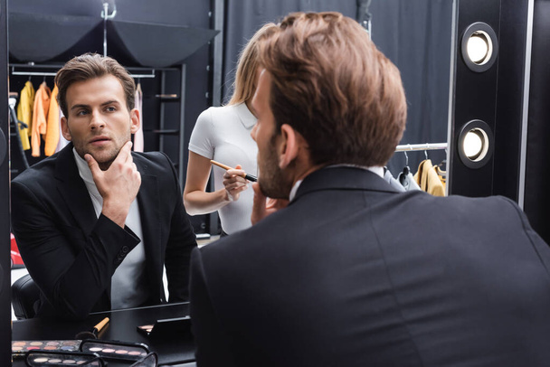 young man touching face while looking in mirror near makeup artist  - Foto, Imagen