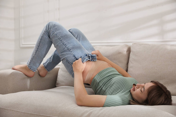 Young woman struggling to squeeze into tight jeans while lying on sofa at home - Fotografie, Obrázek