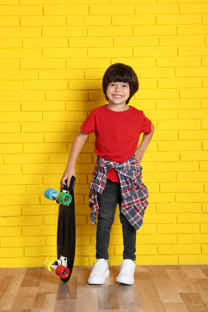 Cute little boy with skateboard near yellow brick wall indoors - Foto, Imagen