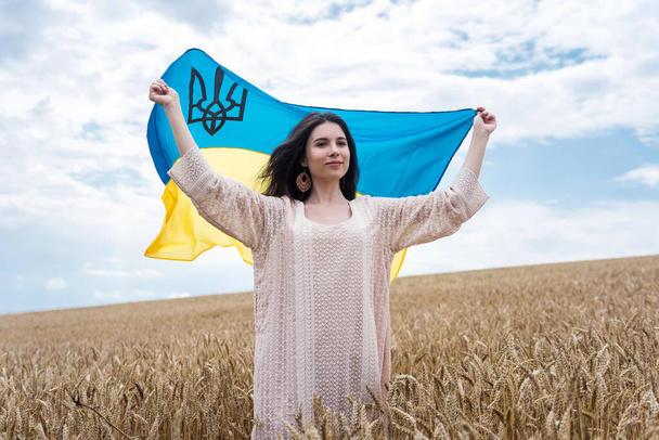 Chica bonita con la bandera de Ucrania pasar tiempo libre en un campo de trigo. Disfrute de un día soleado y caliente - Foto, imagen