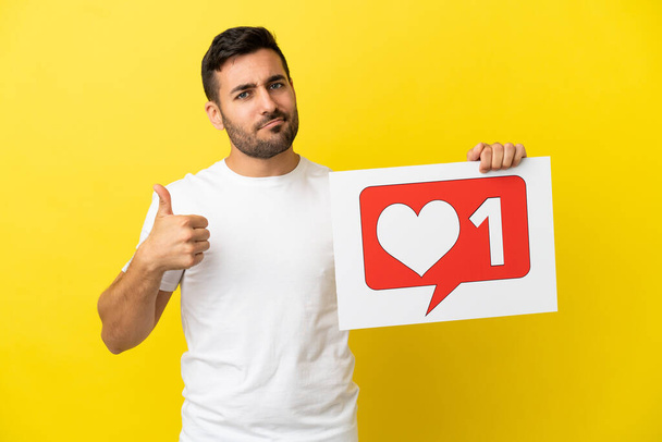 Young handsome caucasian man isolated on yellow background holding a placard with Like icon with thumb up - Foto, Imagem