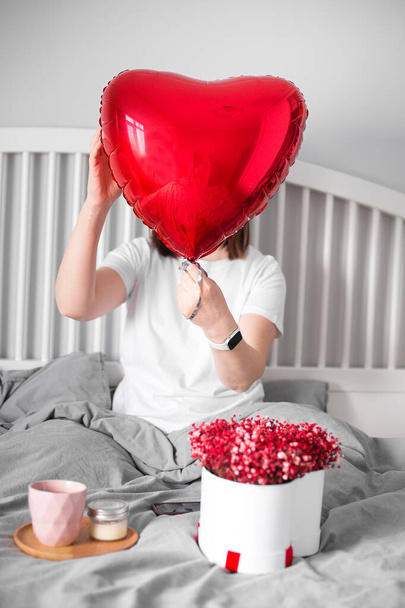 Woman sitting on bed with red baloon in shape of heart and gift box with bouquet gypsophila. Celebrating Saint Valentines Day. Lifestyle concept - Fotó, kép