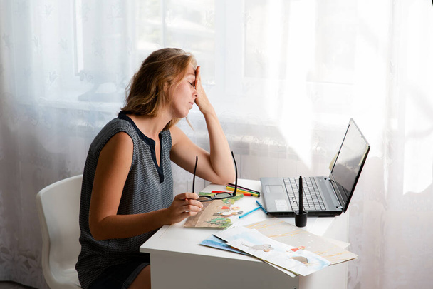 Stress au travail. Une jeune femme assise à son bureau se frotte les yeux fatigués. Sur la table se trouvent un ordinateur portable et des dessins. Espace de copie. Le concept d'auto-isolement et de freelance. - Photo, image