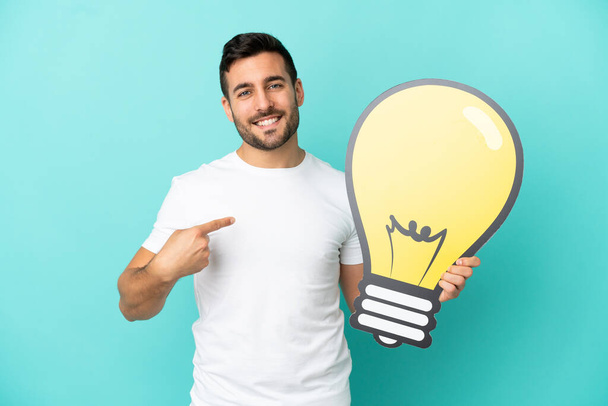 Young handsome caucasian man isolated on blue background holding a bulb icon and pointing it - Fotó, kép
