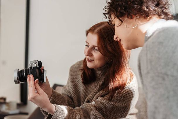 Μια κάθετη φωτογραφία μιας κοκκινομάλλας να τραβάει μια κάμερα και να δείχνει μια φωτογραφία σε μια σγουρή γυναίκα. - Φωτογραφία, εικόνα