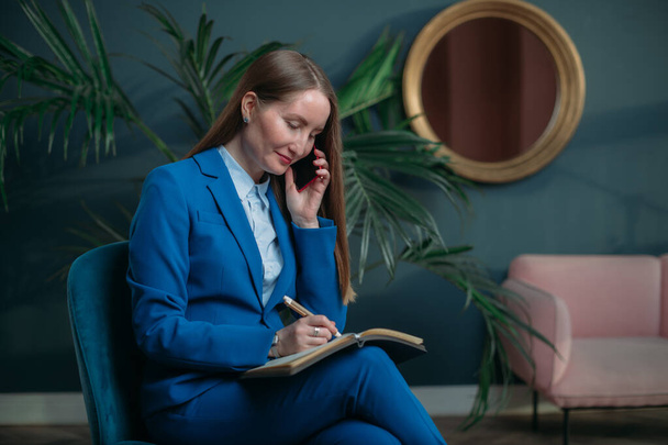Business woman portrait in modern office interior. Stylish young girl in a blue business suit talking on the phone. Woman in business, portrait of a businesswoman. - Foto, afbeelding