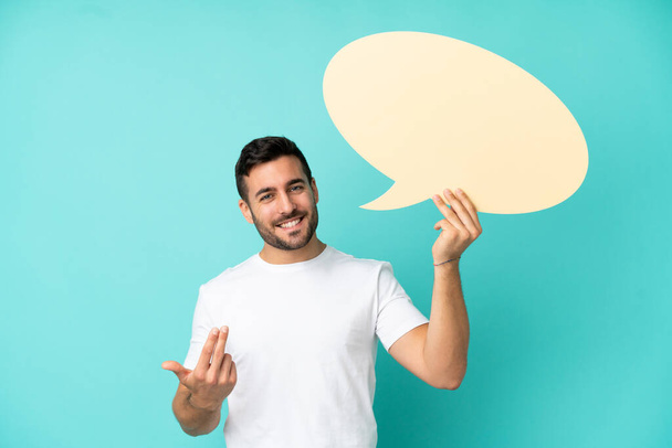 Young handsome caucasian man isolated on blue background holding an empty speech bubble and doing coming gesture - Photo, Image
