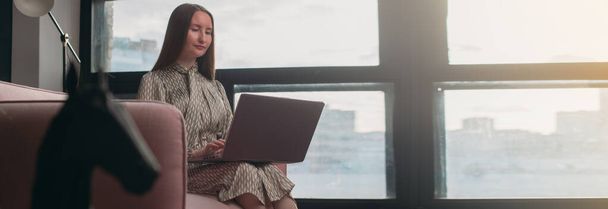 Портрет деловой женщины в современном офисном интерьере с ноутбуком. Стильная улыбающаяся молодая девушка в деловом платье с ноутбуком на диване. Женщина в бизнесе, портрет предпринимательницы. - Фото, изображение
