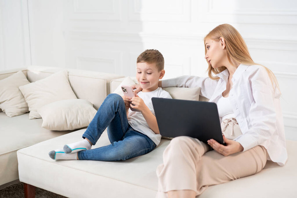 Mamá e hijo en el sofá sentados en el teléfono. Los niños a distancia aprendizaje y las mamás trabajan en el ordenador portátil. - Foto, imagen