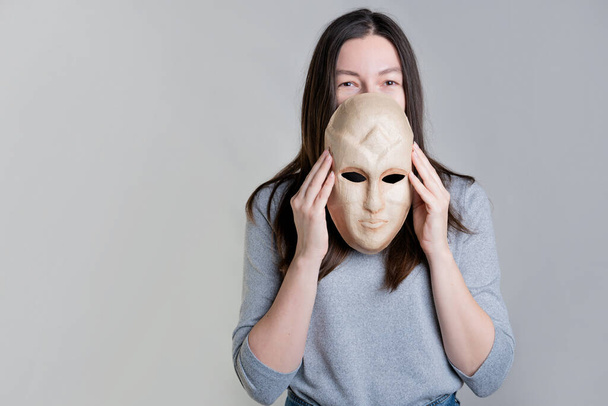 A young woman holds a mask in her hands, hiding her face half, a sly look over the mask. - Valokuva, kuva