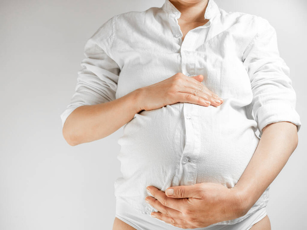 Леди на третьем триместре беременности. Беременная молодая женщина в белой рубашке схватила живот на белом фоне. Девушка на последнем месяце беременности, кладущая руки на живот, обнимает ребенка - Фото, изображение