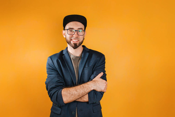 The concept of success and business. A man in a jacket and cap, with a beard and glasses, put his arms around his shoulders and smiled. Orange background. Copy space. - Photo, Image