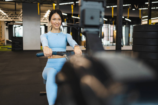 Cheerful woman in blue sportswear on rowing machine. Rowing exercise in the gym - Photo, Image