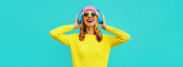 Portrait of happy smiling young woman with headphones listening to music wearing colorful yellow sweater on blue background - Fotografie, Obrázek