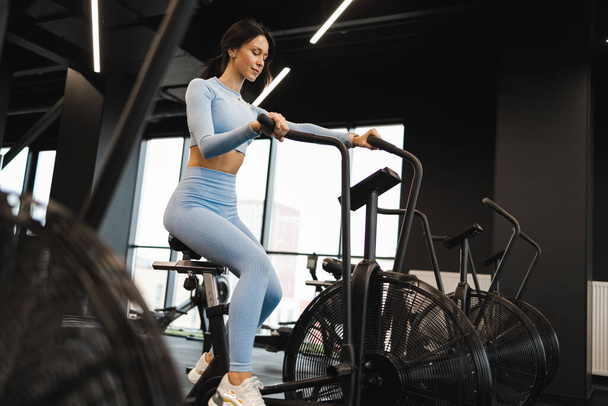 Красивая женщина в кроссфит тренажерный зал с помощью велотренажера для кардио тренировки. Спортивная девушка на воздушном велосипеде - Фото, изображение