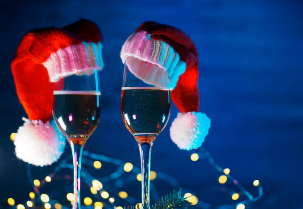Два бокала шампанского в шляпах Санта-Клауса на синем деревянном фоне. Гарландс, Боке - Фото, изображение