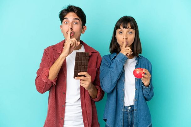 Mladý smíšený závod pár drží jablko a čokoládu izolované na modrém pozadí ukazující znamení mlčení gesto dát prst do úst - Fotografie, Obrázek