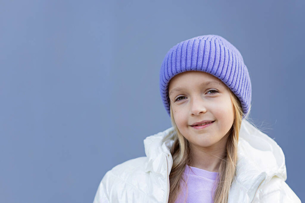 Cute little caucasian girl eight years old with blonde hair smiling outdoor. Kid wearing stylish shirt and knitted hat violet color. Trendy color of the 2022 year very peri - Foto, afbeelding