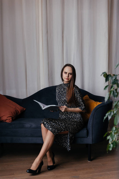 a woman in a dress with polka dots and classic black shoes sits on a dark blue sofa with red and yellow pillows against the background of a beige curtain - Fotó, kép