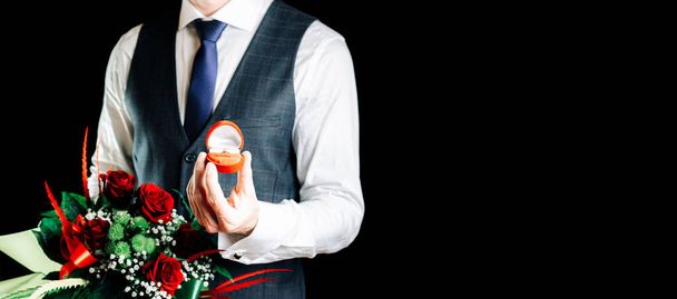 Heiraten Sie mich. Junger glücklicher Mann mit roter Geschenkbox mit diamantenem Verlobungsring, rotem Rosenstrauß. Verlobungsring - Foto, Bild