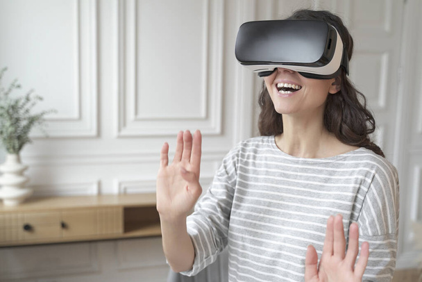 Młoda podekscytowana kobieta w słuchawkach VR interakcji z rozszerzonym światem, grając w gry w domu - Zdjęcie, obraz