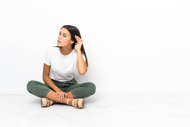 Teenager dívka sedí na podlaze a něco poslouchá tím, že položí ruku na ucho - Fotografie, Obrázek