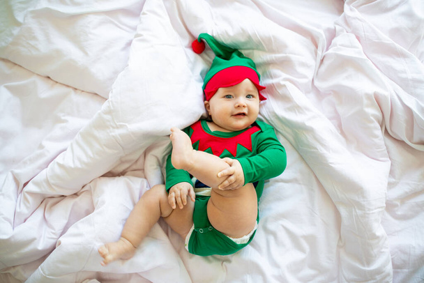 Roztomilá běloška, šest měsíců stará, v elfím kostýmu zelené barvy. Rozkošný Santa pomocník ležící na posteli doma na bílém pozadí - Fotografie, Obrázek