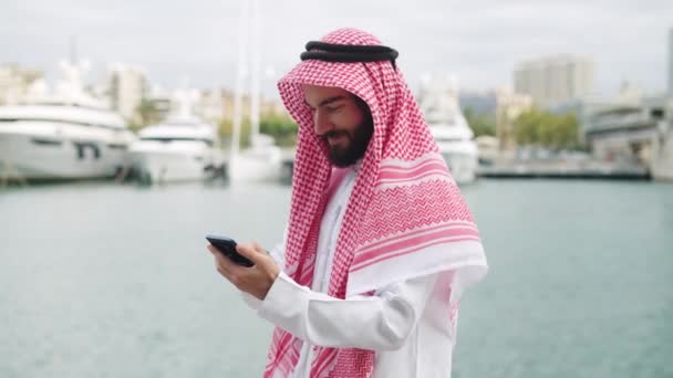 Tradycyjnie ubrany arabski biznesmen ze smartfonem w porcie, czytający wiadomości internetowe przez telefon komórkowy  - Materiał filmowy, wideo