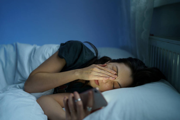 Виснажена жінка лежить у ліжку без сну тримає смартфон, читаючи про розлад сну та психічне здоров'я
 - Фото, зображення