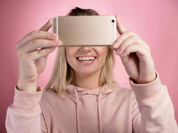 attractive young woman in a pink hoodie uses a smartphone for video communication or photos. - Fotografie, Obrázek