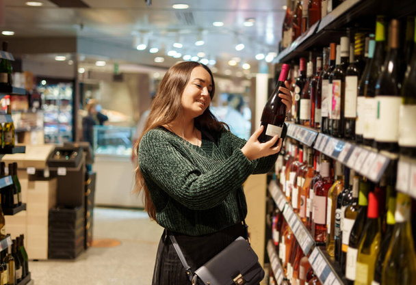 Feliz joven mujer eligiendo vino tinto favorito de pie junto a los estantes en la tienda de comestibles - Foto, Imagen