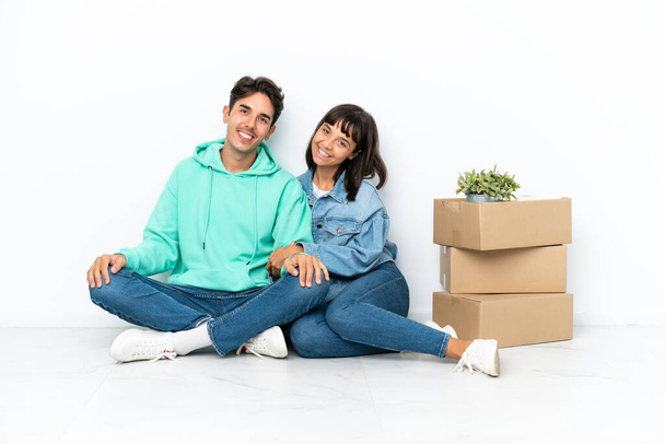 Молодая пара делает шаг, поднимая коробку с вещами, сидя на полу изолированы на белом фоне, держа руки скрещенными в боковом положении, улыбаясь - Фото, изображение