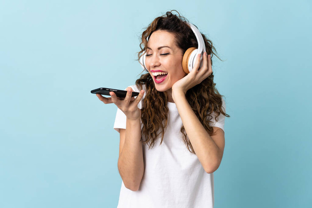 Junge kaukasische Frau isoliert auf blauem Hintergrund Musik hören mit einem Handy und singen - Foto, Bild