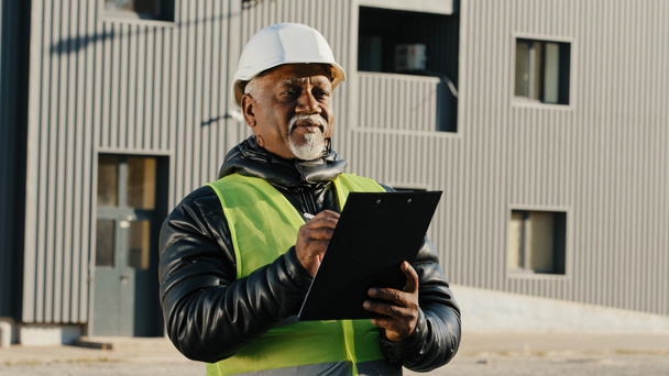 Elderly african american man construction manager evaluates building old foreman standing on street in protective hardhat writes data to tablet engineer builder thinks over work plan closeup outdoors - Photo, Image