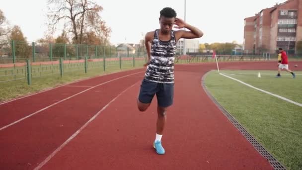 Una joven negra va a practicar deportes en la cinta de correr del estadio y hace ejercicios de estiramiento estáticos. Movimiento lento - Imágenes, Vídeo