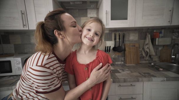 Mutter und kleine Tochter haben gemeinsam Spaß in der Küche. Handeln. Lächelnde Mutter küsst ihr süßes kleines Mädchen auf die Wange auf Küchenmöbeln Hintergrund, Konzept der glücklichen Familie. - Foto, Bild
