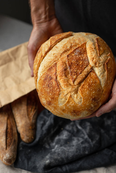 an adult European male baker holds a round fresh bread in his hands. a man in a bakery holds a yeast-free bread on sourdough and a baguette.  - Foto, Imagem