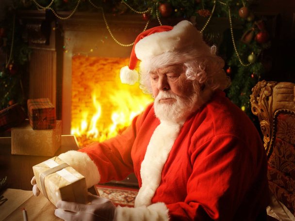 Portrait of happy Santa Claus sitting at his room at home near Christmas tree with gift box - Фото, изображение