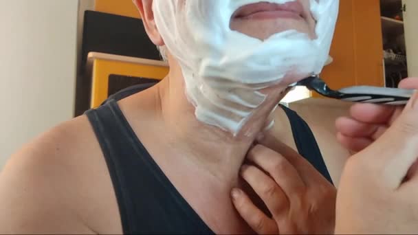 De man scheert zich met een scheermes, verwijdert gezichtsstoppels en overvloedig wit scheerschuim. Video van een deel van het gezicht van een man close-up in een badkamer. - Video