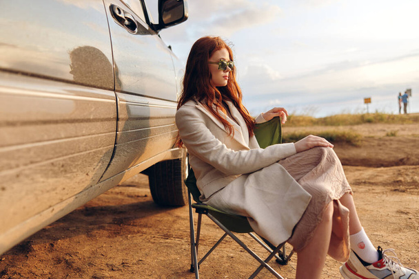 beautiful woman sitting on a chair in a field wearing dark glasses near the car endless field - Photo, image