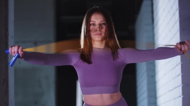 Młoda piękna kobieta w fioletowym stroju sportowym bawi się skakanką w dłoniach i patrzy w kamerę - Materiał filmowy, wideo