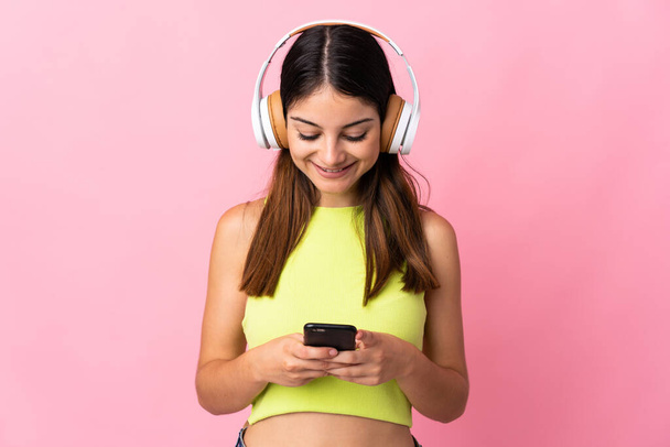 Young caucasian woman isolated on pink background listening music and looking to mobile - Foto, imagen