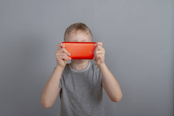 The boy is playing a game on a red smartphone and has covered his face and eyes with the gadget screen. - Foto, imagen