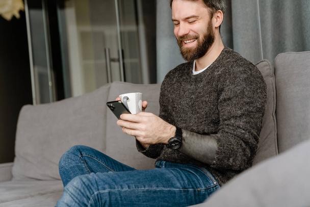 Baard man met behulp van mobiele telefoon en het drinken van koffie tijdens het zitten op de bank thuis - Foto, afbeelding